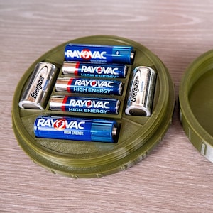 May include: A green plastic container with a lid, designed to hold batteries. The container has six compartments, each holding a different type of battery, including Rayovac and Energizer brands.