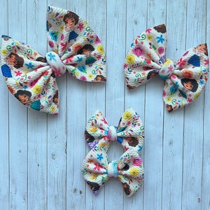 May include: Four fabric hair bows in varying sizes. The bows are cream-colored with a repeating pattern of a cartoon character, flowers, and leaves. The bows are tied in the center with a matching fabric.