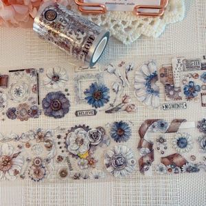 Puede incluir: Un rollo de cinta adhesiva transparente con un patrón repetitivo de flores azules y marrones, engranajes mecánicos, marcos de estilo vintage y etiquetas de texto que incluyen "BELIEVE", "MOMENTS" y "TOGETHER".