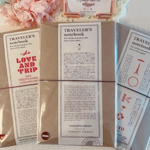 May include: Three Traveler's Company notebooks in brown leather covers, each wrapped in a paper band with text and graphics. The center notebook is labeled Starbucks Reserve. A rose gold metal stand holds a Moondreamer Studio business card.