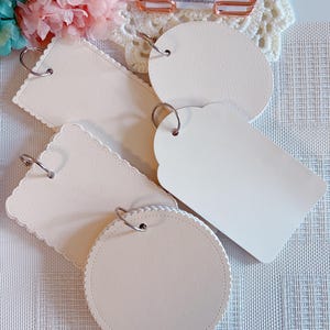 May include: A selection of cream-coloured blank tags in diverse shapes, including round, scalloped, and rectangular. Each tag features a metal ring for attachment. The tags are displayed on a textured surface, with flowers in the background. The text "Moondreamer STUDIO" is visible.