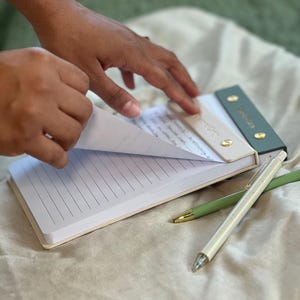 Può includere: Un blocco note con copertina color crema e pagine a righe viene aperto. Il blocco note ha una parte superiore verde e crema con dettagli dorati. Due penne, una color crema e una verde, sono accanto al blocco note.