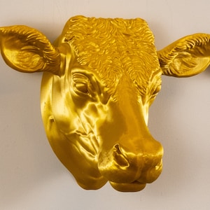 May include: A golden cow's head sculpture with detailed features, including textured fur and a realistic expression. The sculpture is mounted on a white wall.