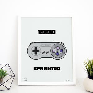 May include: A white framed print with a gray background featuring a black and white illustration of a Super Nintendo controller with the text "1990" and "SPR NNTODO" in black. The controller has the text "SUPER NOODLES" printed on it.