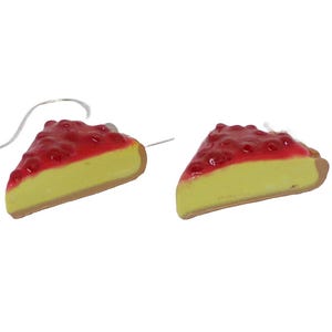Può includere: Orecchini a forma di fette di cheesecake. Ogni orecchino presenta una base di torta gialla, una crosta marrone e una glassa rossa con frutti di bosco. Gli orecchini hanno fili argentati.