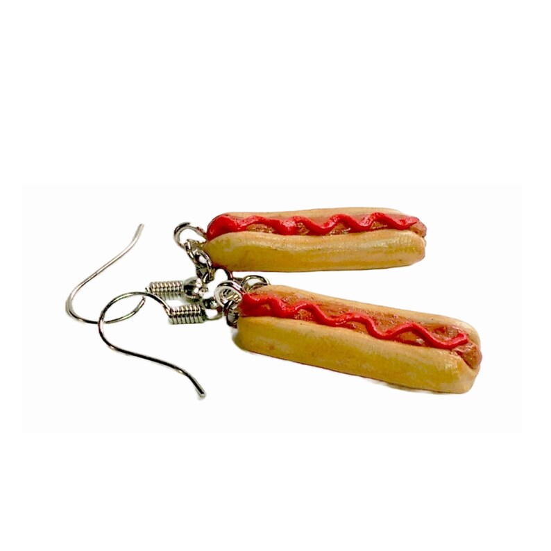 May include: A pair of dangle earrings featuring a miniature hot dog with ketchup on a bun. The earrings are made of polymer clay and have silver hooks.