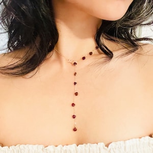 May include: A delicate gold chain necklace with small, dark red gemstone beads. The necklace features a Y-shaped design with a longer strand extending down the center.