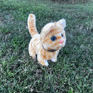 May include: A plush orange and white tabby cat toy with blue eyes and a black and white polka dot collar. The toy is sitting on a bed of green grass.