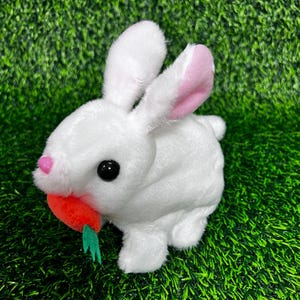 May include: A white plush bunny toy with pink inner ears, a pink nose, and black eyes. The bunny is holding a plush orange carrot with green leaves. The toy is set against a green background.