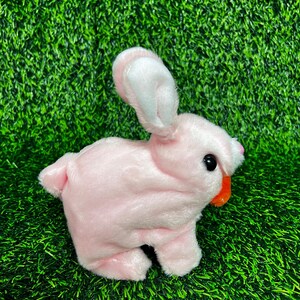 May include: A pink plush bunny toy with black eyes and an orange mouth. The bunny has long ears with pink inner ears and is posed on a green surface.