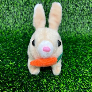 May include: A plush toy bunny with beige fur, black eyes, and a pink nose. The bunny is holding an orange carrot with a green leaf. The toy is set against a backdrop of green artificial grass.