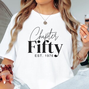 May include: White t-shirt with the text "Chapter Fifty EST. 1976" in black script and bold font. The shirt is a casual style, suitable for everyday wear. The design celebrates a milestone birthday.