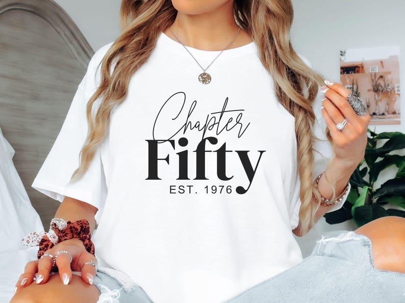 May include: White t-shirt with the text "Chapter Fifty EST. 1976" in black script and bold font. The shirt is a casual style, suitable for everyday wear. The design celebrates a milestone birthday.