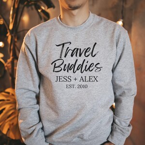 May include: Gray sweatshirt with the text "Travel Buddies" in black script font. Below the text is "Jess + Alex Est. 2010" in black.