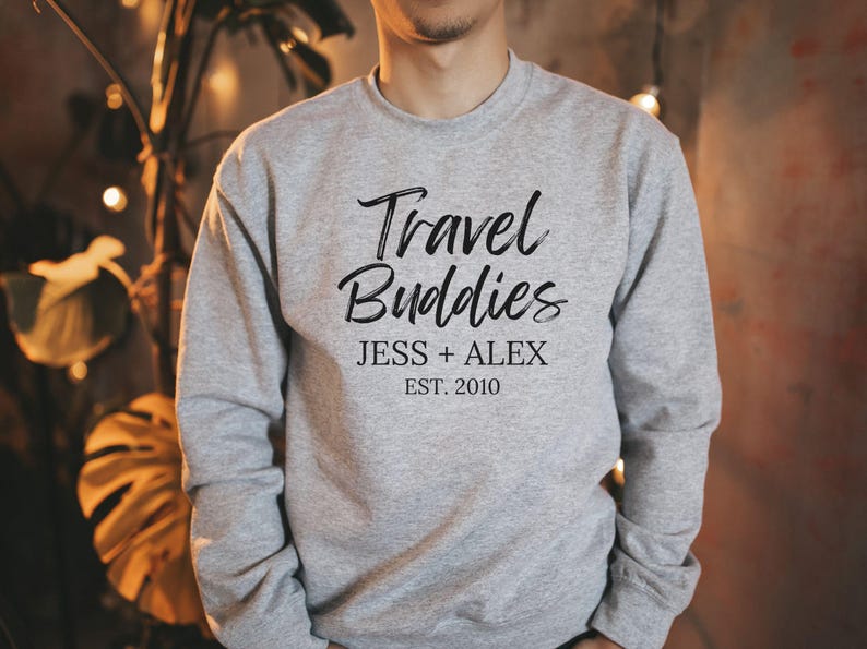 May include: Gray sweatshirt with the text "Travel Buddies" in black script font. Below the text is "Jess + Alex Est. 2010" in black.