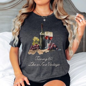 Pode incluir: Uma camiseta cinza escura com um gr&aacute;fico de uma garrafa de vinho e duas ta&ccedil;as de vinho. O texto "Turning 60, Like a Fine Vintage" est&aacute; impresso na camiseta.