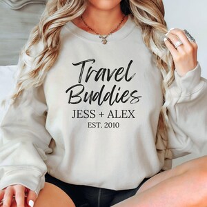 May include: A white sweatshirt with the text "Travel Buddies" in black script font. Below the text is "Jess + Alex Est. 2010" in black.