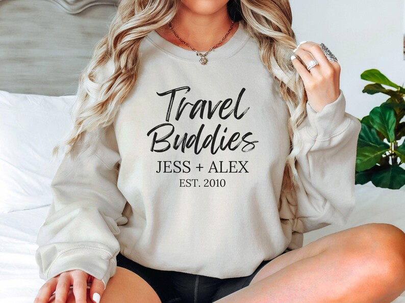 May include: A white sweatshirt with the text "Travel Buddies" in black script font. Below the text is "Jess + Alex Est. 2010" in black.