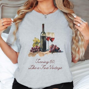 Pode incluir: Camiseta cinza com uma ilustra&ccedil;&atilde;o em aquarela de uma garrafa de vinho, duas ta&ccedil;as de vinho e uvas. O texto "Turning 60, Like a Fine Vintage" est&aacute; impresso na camiseta.