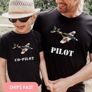 May include: A black t-shirt with a camouflage airplane graphic and the text "Pilot" on the front. A smaller black t-shirt with the same airplane graphic and the text "Co-Pilot" on the front. The shirts are being worn by a father and son.