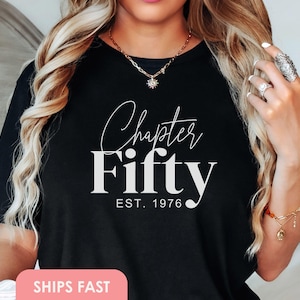 May include: Black t-shirt with the words "Chapter Fifty" in white script and block letters, with "EST. 1976" below. The shirt is a casual style, suitable for milestone birthdays. The image shows the shirt on a person.