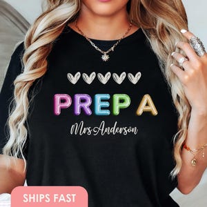 Könnte beinhalten: Schwarzes T-Shirt mit dem Wort "PREPA" in bunten Buchstaben und dem Namen "Mrs Anderson" in Schreibschrift darunter. Fünf weiße Herzen befinden sich über dem Wort "PREPA".