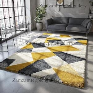 May include: A rectangular area rug with a geometric pattern of yellow, gray, and white triangles. The rug is placed on a concrete floor in a living room setting, with a gray sofa and large windows in the background.