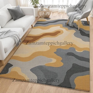 May include: A rectangular area rug with a layered, abstract design in shades of gray, tan, and gold. The rug features a soft, textured surface and is placed on a hardwood floor. The text "Zamzamteppichgallery" is printed on the rug.