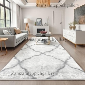 May include: A large, rectangular area rug with a light gray and white geometric pattern dominates the room. The rug's design features a repeating quatrefoil motif. The room includes a gray sofa, a black coffee table, and a white cabinet. The text "Zamzamteppichgallery" is visible.