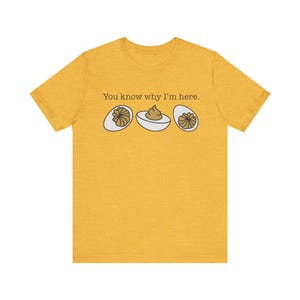 Op de afbeelding: Mosterdgeel T-shirt met de tekst "You know why I'm here." en een afbeelding van drie gevulde eieren. De eieren zijn wit met een bruine vulling en een decoratieve garnering.