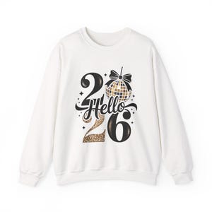Puede incluir: Sudadera blanca con el texto "Hello 26" en negro y purpurina dorada. El diseño incluye una bola de discoteca y un lazo, ideal para una celebración de Año Nuevo.