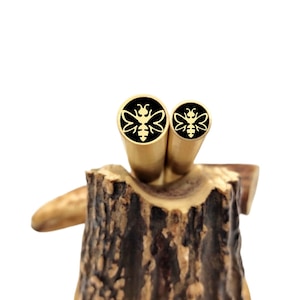 Könnte beinhalten: Zwei goldfarbene Metall-Stiftrohlinge mit schwarzem Hintergrund und einem Bienendesign. Die Rohlinge liegen auf einem Stück Holz.