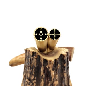 May include: Two gold-colored metal pen blanks with black crosshair targets on the ends. The blanks are sitting on a piece of wood.