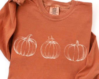 May include: Rust-colored long-sleeve shirt with a crew neck. The shirt features a white line drawing of three pumpkins across the front. The lower left corner has a small orange circle with the word "YAM" printed on it.