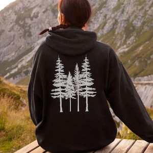 Könnte beinhalten: Ein schwarzer Kapuzenpullover mit einer weißen Silhouette von drei Kiefern auf der Rückseite. Die Person, die den Pullover trägt, steht vor einer Bergkette.