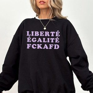 May include: Black sweatshirt with the words "LIBERTÉ ÉGALITÉ FCKAFD" in lavender lettering. The sweatshirt has a crew neck and long sleeves. The overall style is casual and comfortable.