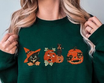 May include: Dark green sweatshirt featuring a Halloween-themed design. The design includes a black cat wearing a witch hat, pumpkins, and a black cat inside a pumpkin. The sweatshirt is a crew neck style.