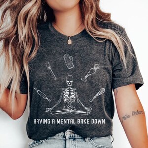 Könnte beinhalten: Ein dunkelgraues T-Shirt mit einem weißen Skelett-Design, das mit Backutensilien um sich herum meditiert. Der Text "HAVING A MENTAL BAKE DOWN" ist unter dem Skelett aufgedruckt. Das Model trägt eine blaue Jeans und weiße Turnschuhe.