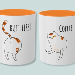 May include: Two white coffee mugs with orange rims. One mug features a cartoon cat with the text "Butt First" and the other mug features a cartoon cat with the text "Coffee".