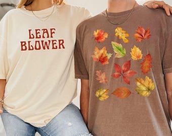 May include: Two short-sleeved t-shirts. One is cream-colored with the text "LEAF BLOWER" in red. The other is brown and features a collection of colorful autumn leaves. Both shirts are worn by people.