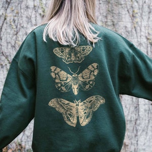 Könnte beinhalten: Ein dunkelgrüner Pullover mit einem goldenen Aufdruck von drei Motten auf der Rückseite.
