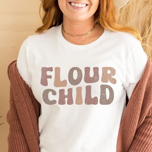 Könnte beinhalten: Weißes T-Shirt mit dem Text "Flour Child" in einer retro-farbigen Schrift. Das Shirt wird von einer Person mit einem Cardigan getragen.