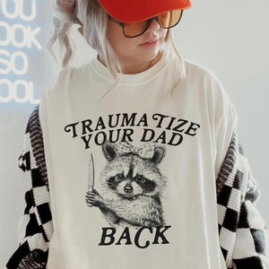May include: A white t-shirt with the text "TRAUMATIZE YOUR DAD BACK" and a raccoon graphic holding a knife. The person is wearing a red cap with "MAD LOVE FOR YOU" and a black and white checkered cardigan.