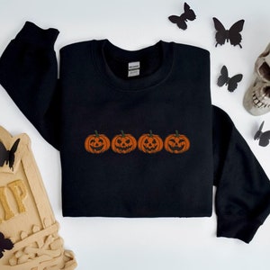 Könnte beinhalten: Schwarzes Sweatshirt mit einem gestickten Design von vier orangefarbenen Kürbissen mit geschnitzten Gesichtern. Die Kürbisse sind in einer Reihe über der Brust angeordnet. Das Sweatshirt ist ein Rundhals-Stil.