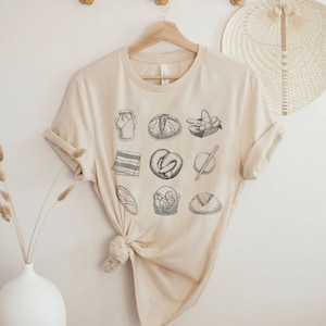 May include: A beige t-shirt with a black and white graphic print of various types of bread, including a loaf, a pretzel, and a baguette. The graphic is arranged in a grid pattern.