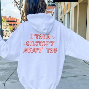 Könnte beinhalten: Weißer Kapuzenpullover mit dem Text "I TOLD CHATGPT ABOUT YOU" in Orange. Der Kapuzenpullover hat eine Kapuze und lange Ärmel. Die Person trägt weiße Turnschuhe und Socken.