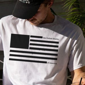 Peut inclure: T-shirt blanc avec un drapeau am&eacute;ricain noir et blanc et le texte "the United States, of America." Une personne porte une casquette noire et tient une boisson au caf&eacute;.