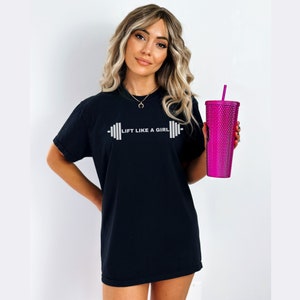 K&ouml;nnte beinhalten: Schwarzes T-Shirt mit dem Text "Lift Like A Girl" und einer Hantelgrafik. Die Person h&auml;lt einen rosa Becher mit Strohhalm.