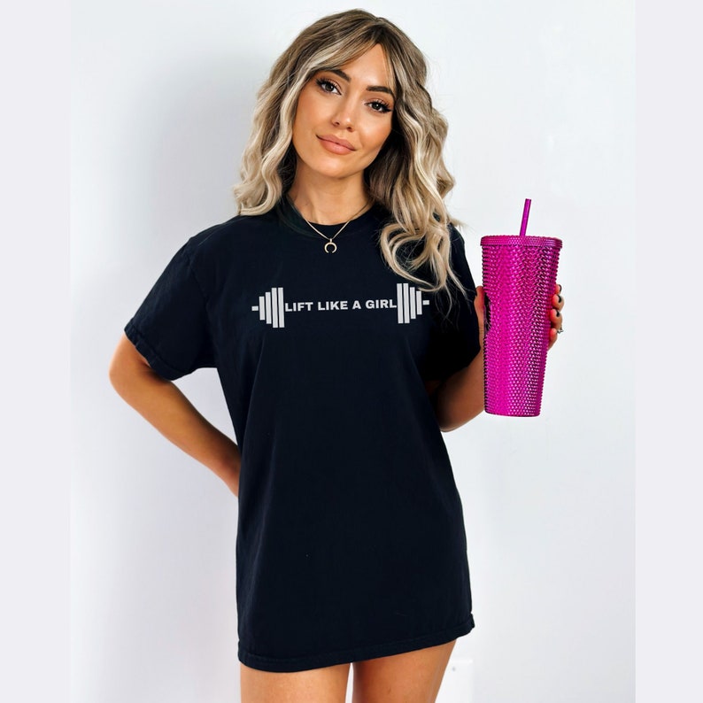 K&ouml;nnte beinhalten: Schwarzes T-Shirt mit dem Text "Lift Like A Girl" und einer Hantelgrafik. Die Person h&auml;lt einen rosa Becher mit Strohhalm.