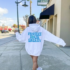 K&ouml;nnte beinhalten: Wei&szlig;er Kapuzenpullover mit einem blauen Augen-Motiv und dem Text "PROTECT YOUR ENERGY" auf der R&uuml;ckseite.